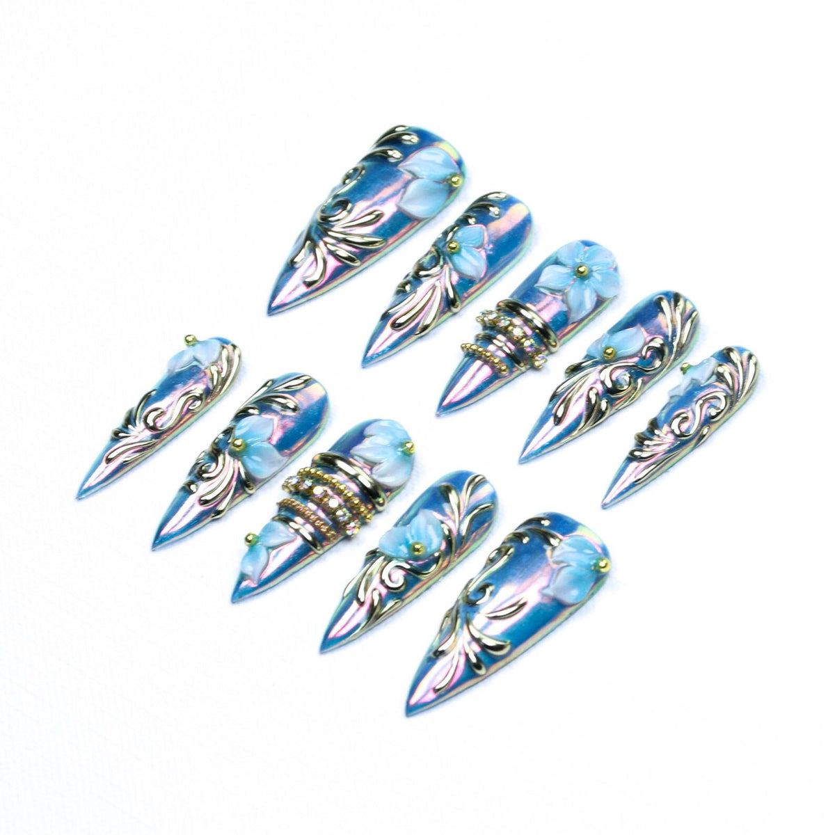 A set of ten stiletto-shaped press-on nails featuring an iridescent blue finish. Each nail is decorated with raised light blue floral accents, silver metallic scrollwork, and small gold studs. Some nails include bands of embedded clear rhinestones.
