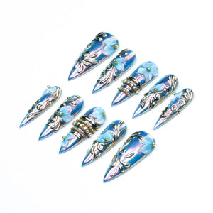 A set of ten stiletto-shaped press-on nails featuring an iridescent blue finish. Each nail is decorated with raised light blue floral accents, silver metallic scrollwork, and small gold studs. Some nails include bands of embedded clear rhinestones.