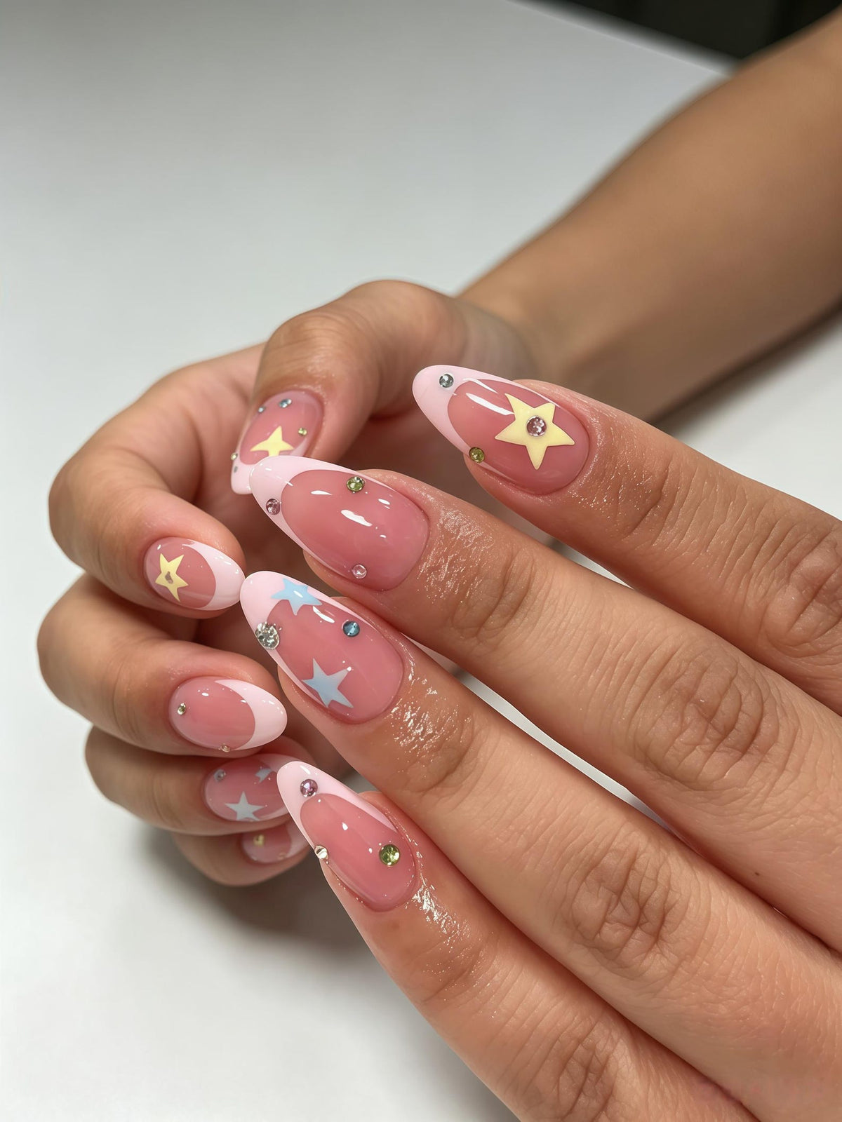 Almond-shaped nails featuring a pink French manicure base with pastel yellow and light blue star decals. The nail art is accented with small, multicolored rhinestones placed individually across the nail beds and along the tips.