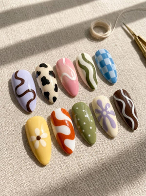 A set of ten almond-shaped press-on nails featuring various designs including a light purple wavy line, black and cream cow print, pink with white swirls, olive green waves, blue checkerboard, yellow daisy, orange swirls, green with white dots, purpl