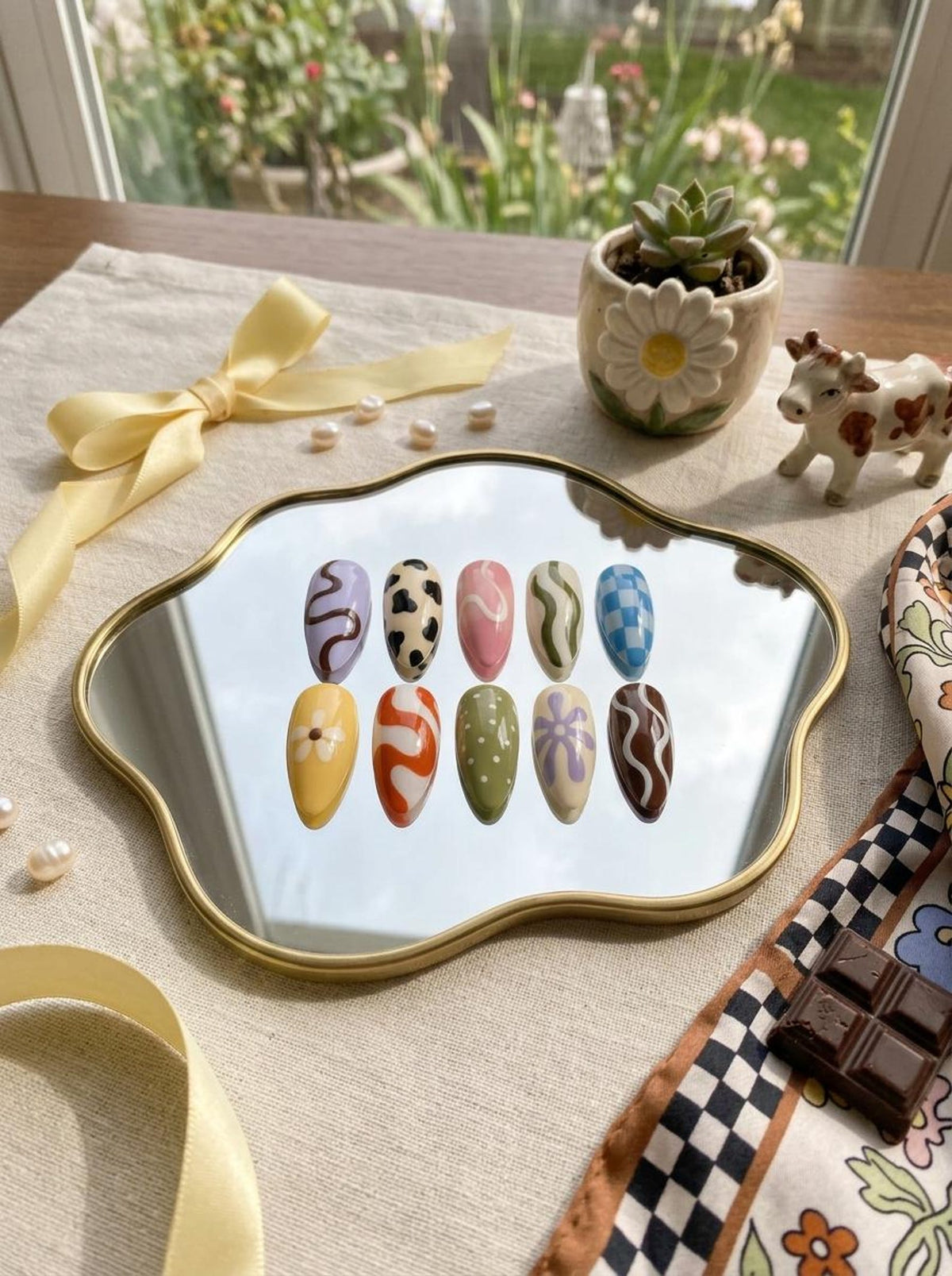 A set of ten almond-shaped press-on nails displayed on a gold-rimmed wavy mirror. The nails feature various designs including cow print, blue and white checkered patterns, white flowers on yellow, green with white dots, and wavy lines.