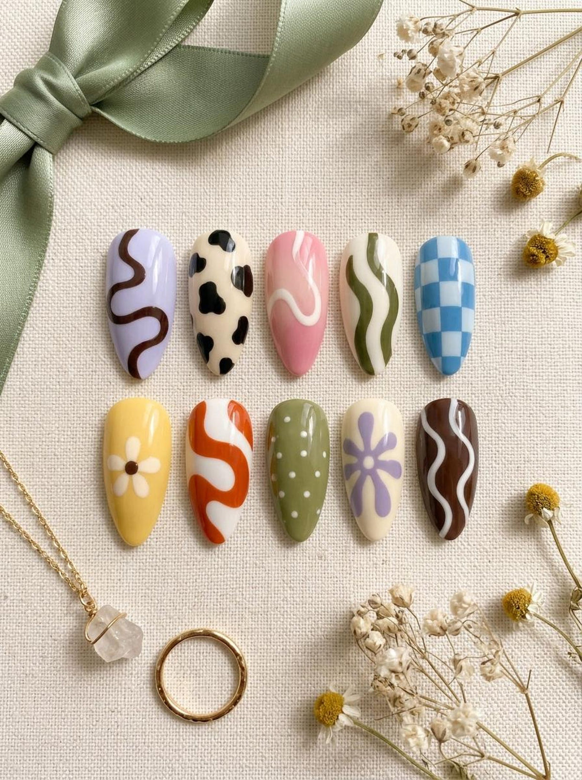 Ten almond-shaped press-on nails featuring diverse designs including cow print, blue and white checkerboard, wavy lines, white polka dots, and floral patterns in colors like lavender, pink, olive green, yellow, orange, and brown.