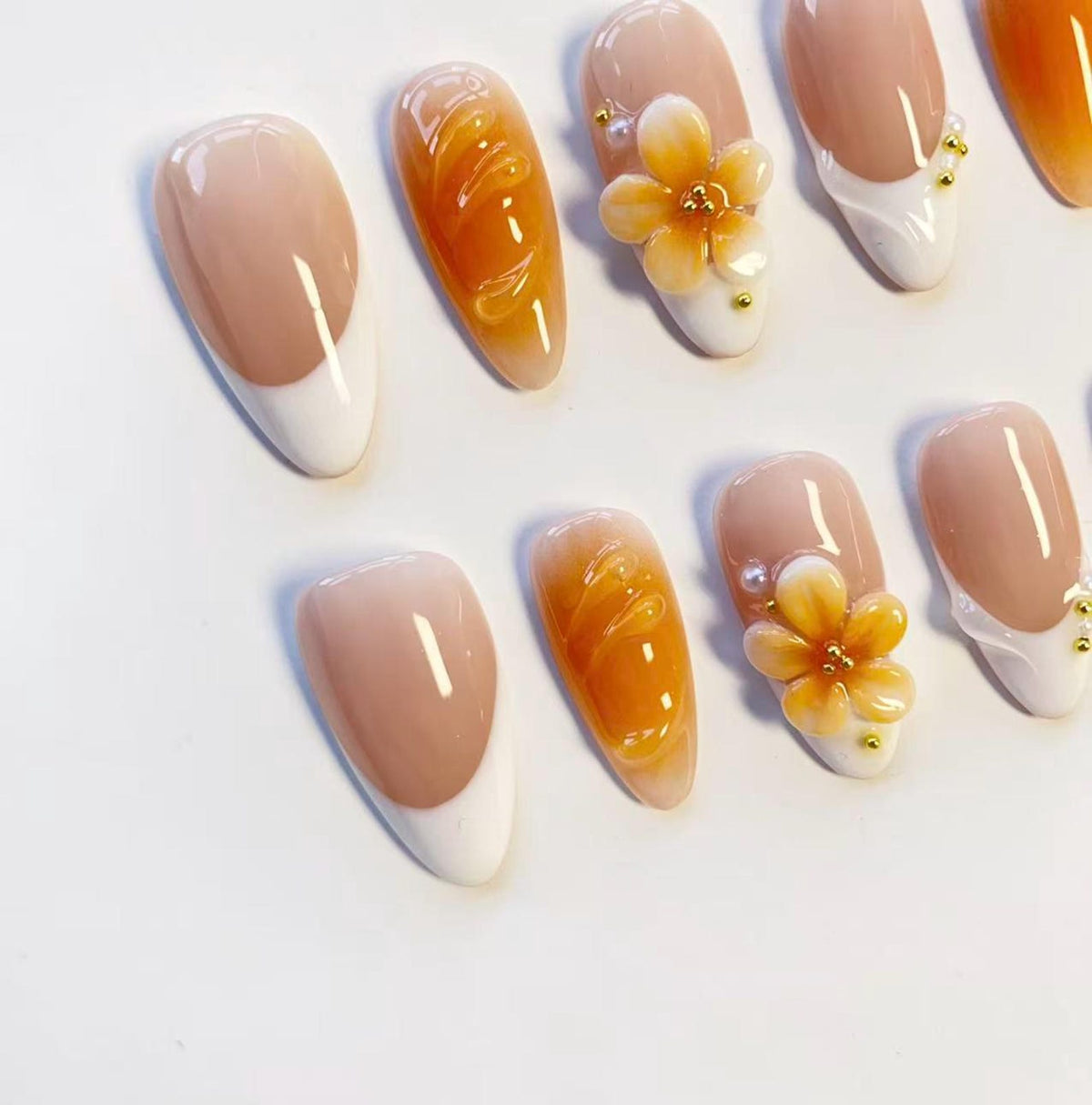 A set of almond-shaped press-on nails featuring a variety of designs including classic white French tips, solid amber-colored nails with raised swirl textures, and nails adorned with three-dimensional orange flower embellishments.