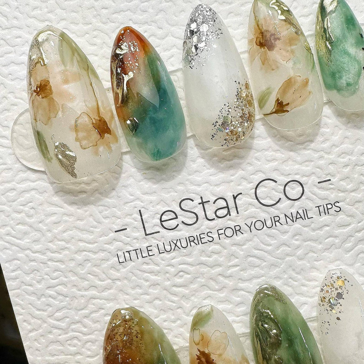 A set of almond-shaped press-on nail tips featuring watercolor floral patterns, green and orange swirls, and silver glitter accents. The display card includes the text - LeStar Co - LITTLE LUXURIES FO
