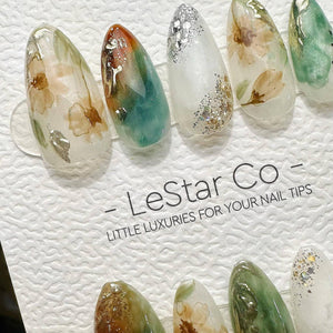 A set of almond-shaped press-on nail tips featuring watercolor floral patterns, green and orange swirls, and silver glitter accents. The display card includes the text - LeStar Co - LITTLE LUXURIES FO