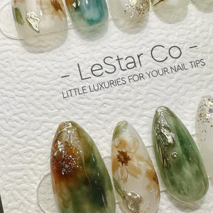 A set of press-on nail tips featuring marbled green, brown, and white patterns with gold foil accents. The display card includes the text LeStar Co and LITTLE LUXURIES FOR YOUR NAIL TIPS printed in bl