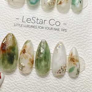 A set of press-on nails featuring a mix of green, white, and gold designs with floral patterns and glitter accents. The display card includes the text LeStar Co - LITTLE LUXURIES FOR YOUR NAIL TIPS. T