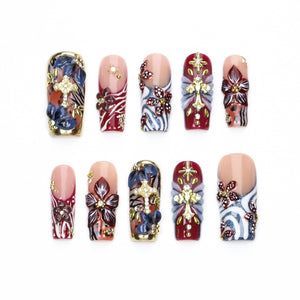 A set of ten press-on nails featuring a nude base with intricate designs including dark blue and burgundy floral patterns, gold cross charms, silver glitter accents, and small clear rhinestones arranged in various decorative motifs.