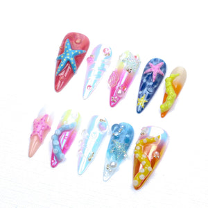 A set of ten stiletto-shaped press-on nails featuring various ocean-themed designs. The nails display gradients of pink, blue, and yellow, decorated with three-dimensional starfish, tentacles, pearls, and iridescent accents.