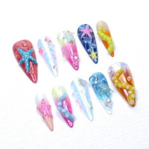 A set of ten press-on nails featuring an ocean theme with various designs including 3D starfish, tentacles, iridescent finishes, and small pearl accents. The nails display a vibrant color palette of pink, blue, yellow, and orange.