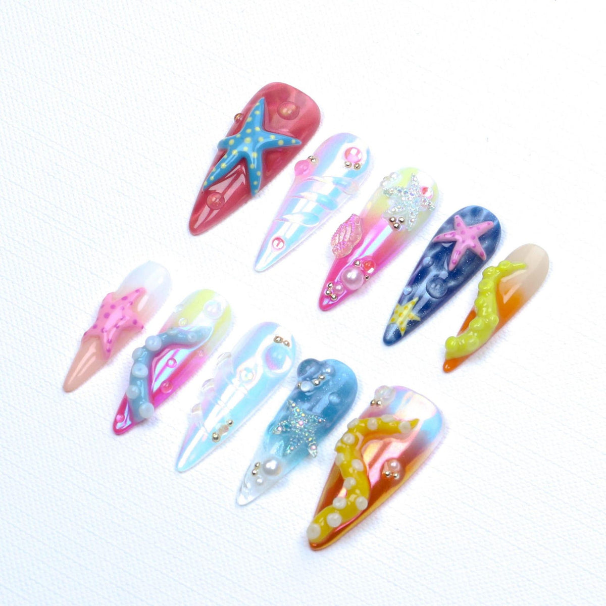 A set of ten stiletto-shaped press-on nails featuring ocean-themed 3D decorations. The designs include colorful starfish, octopus tentacles, pearls, and iridescent finishes in shades of pink, blue, yellow, and white with glossy textures.