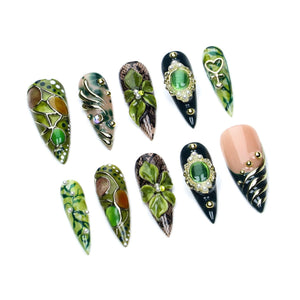 A set of ten press-on nails featuring various designs including green and brown stained-glass patterns, 3D green floral accents, black lace textures, gold metallic swirls, and oval green cabochon stones surrounded by small white pearls.