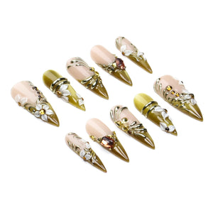 A set of ten stiletto-shaped press-on nails featuring a gradient of nude to olive green. The designs include raised white floral accents, metallic gold swirls, and embedded amber-colored rhinestones across the pointed nail surfaces.