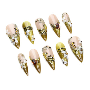 A set of ten stiletto-shaped press-on nails featuring a nude and olive green gradient base. The nails are decorated with white floral accents, gold metallic swirls, and small amber-colored rhinestones arranged in various patterns.