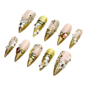 A set of ten stiletto-shaped press-on nails featuring a nude base with olive green tips. The nails are decorated with raised white floral accents, metallic gold swirls, and small amber-colored rhinestones arranged in various patterns.