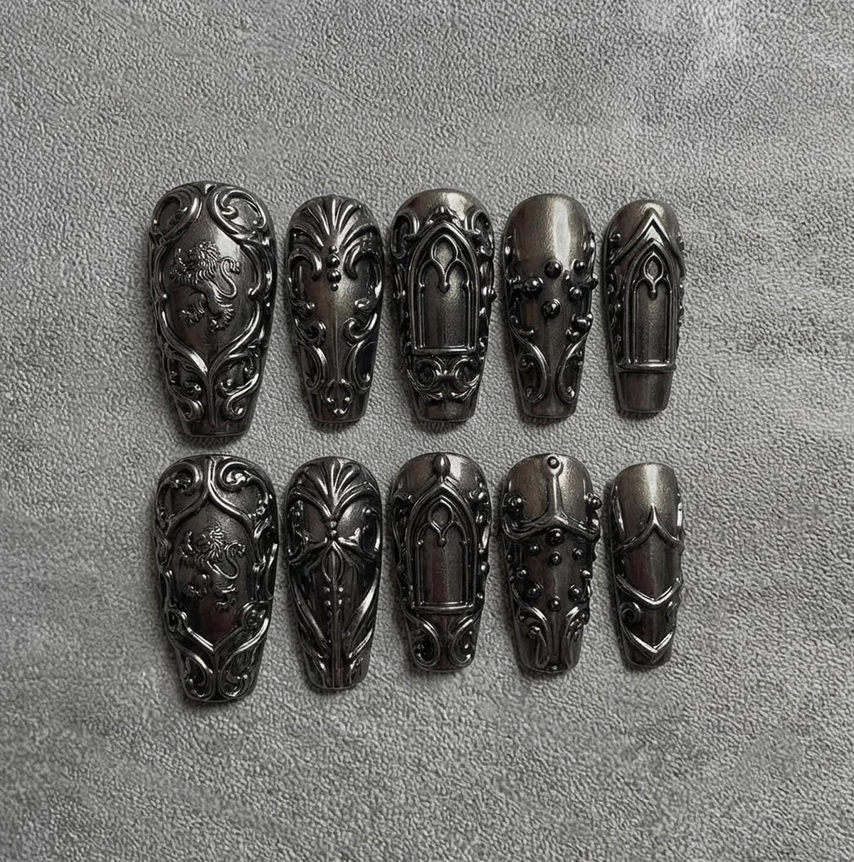A set of ten press-on nails in a dark metallic gunmetal gray finish. Each nail features intricate raised relief designs, including lion motifs, ornate scrollwork, arched window patterns, and small spherical studs in a matching dark tone.