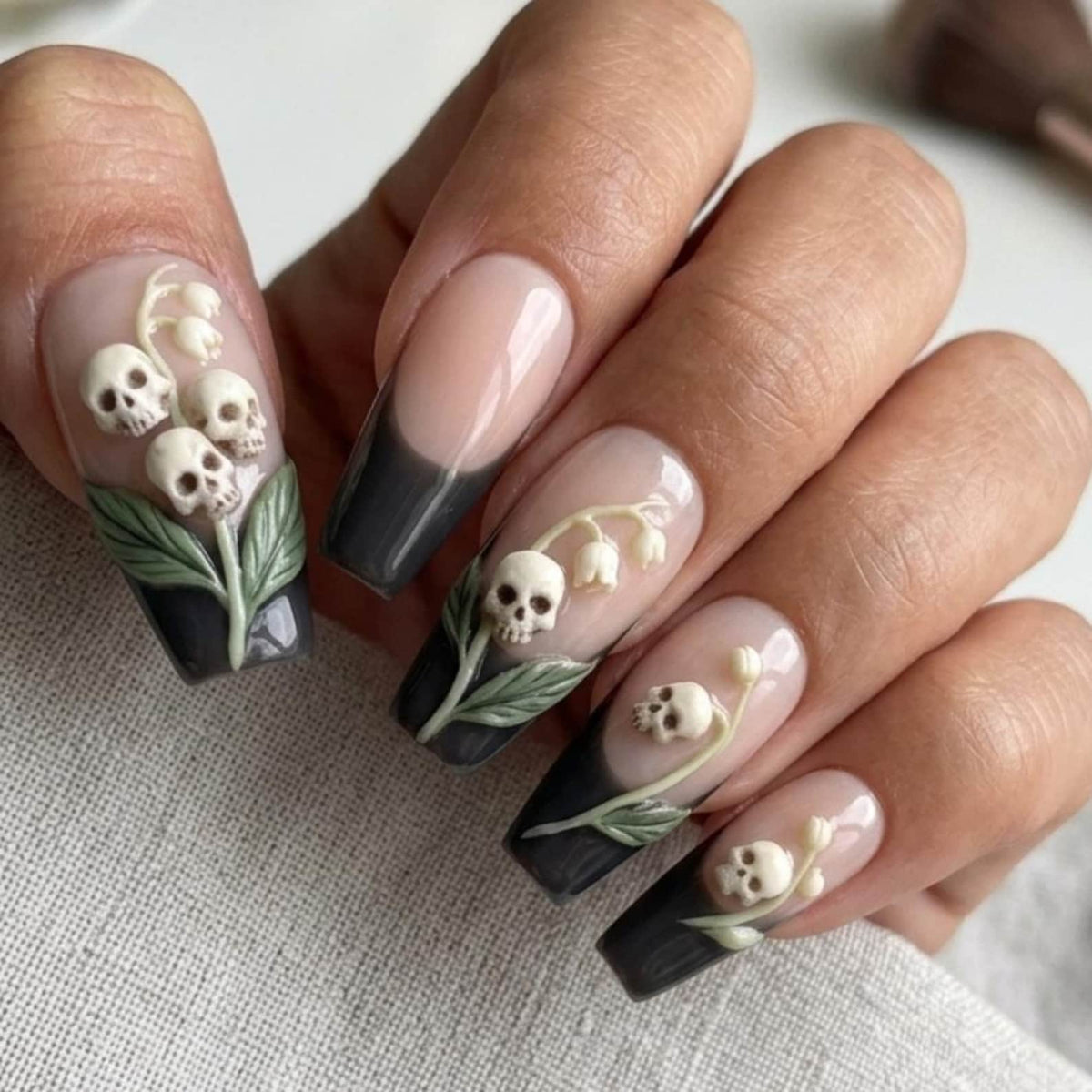 Long coffin-shaped nails featuring a French manicure style with black tips and a nude base. Each nail is decorated with three-dimensional relief art depicting small off-white skulls integrated into stems of lily of the valley flowers.