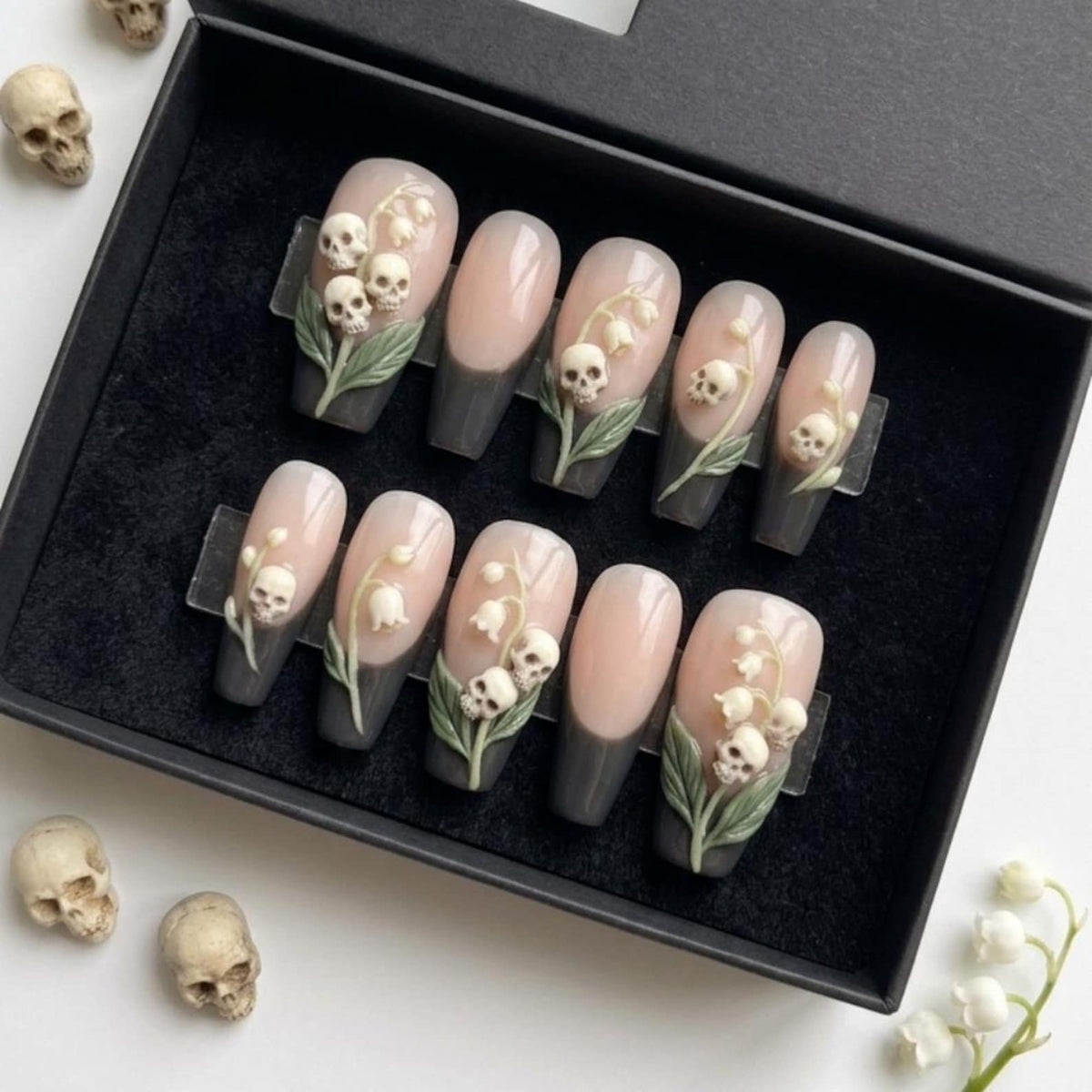 A set of ten press-on nails in a black box, featuring a French manicure style with a dark gray base and nude tips. Several nails are decorated with three-dimensional relief designs of white skulls integrated into lily of the valley flowers.