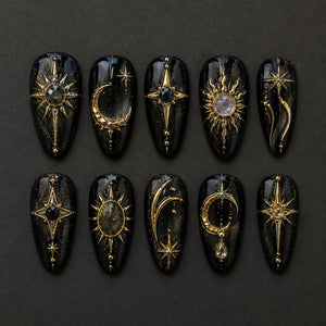 A set of ten almond-shaped press-on nails with a black base featuring gold celestial motifs. Each nail displays unique designs including suns, moons, and stars, accented with small embedded crystals and metallic gold detailing.