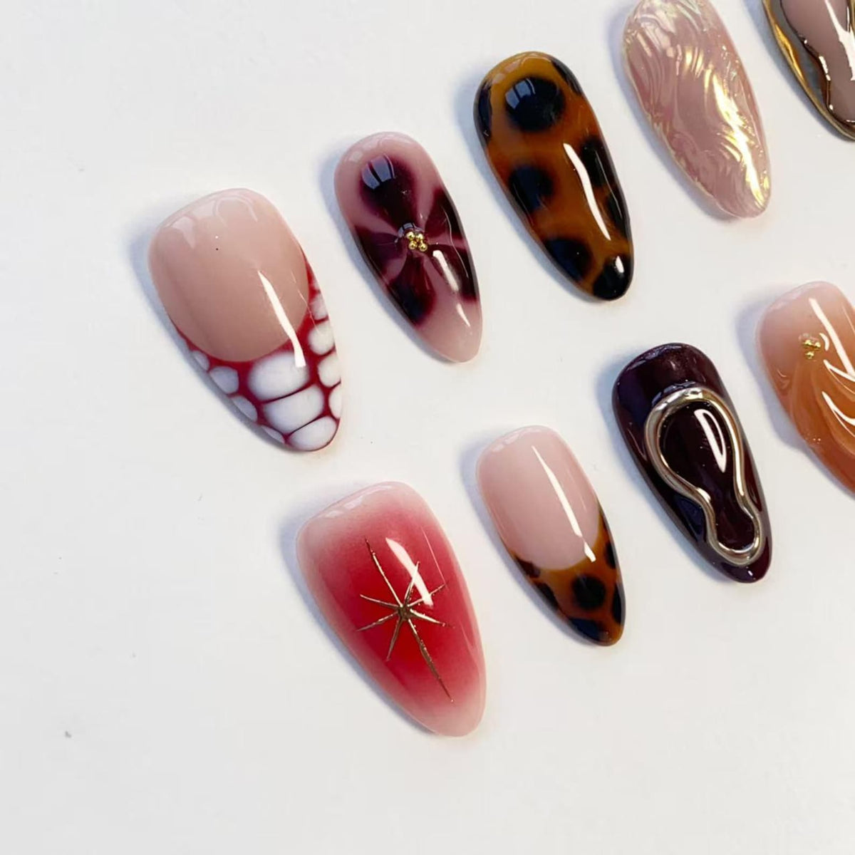 A set of almond-shaped press-on nails featuring diverse designs including a red and white grid pattern, a dark floral motif, brown leopard print, iridescent swirls, a deep burgundy base with gold accents, and a red gradient with a gold star.