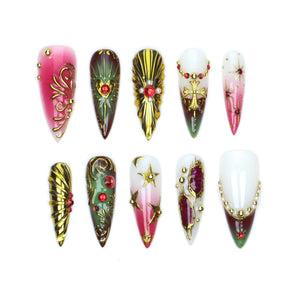 A set of ten stiletto-shaped press-on nails featuring a variety of designs including gold filigree, metallic gold accents, red rhinestones, white and pink gradients, star motifs, and cross emblems arranged in two rows of five.