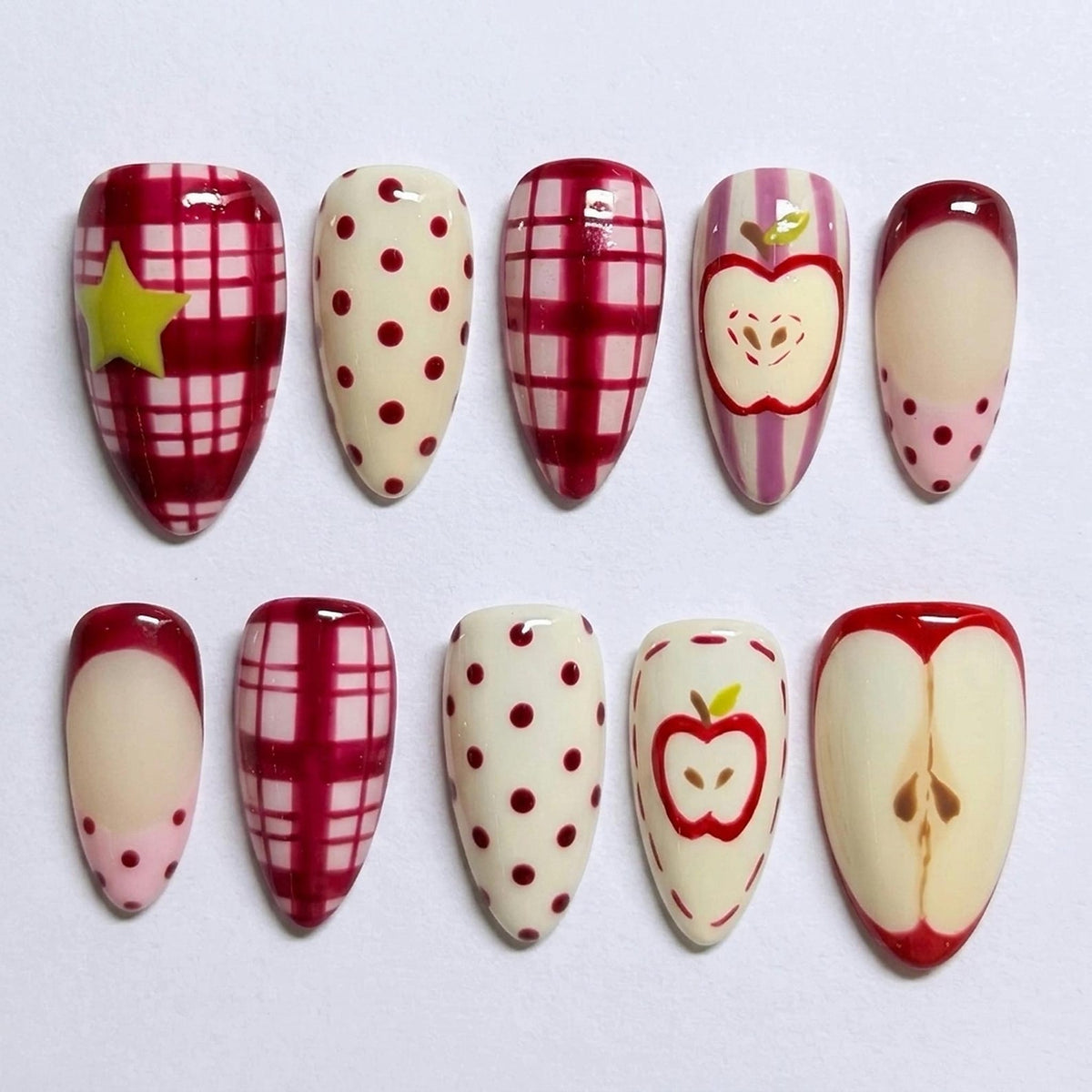 A set of ten press-on nails featuring various designs including red and white plaid patterns, cream backgrounds with red polka dots, a yellow star accent, vertical purple stripes with apple illustrations, and a cross-section of an apple.