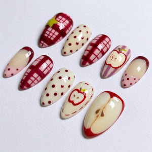 A set of ten almond-shaped press-on nails featuring a variety of designs including red and white plaid patterns, cream-colored bases with red polka dots, a yellow star accent, and detailed illustrations of sliced red apples.