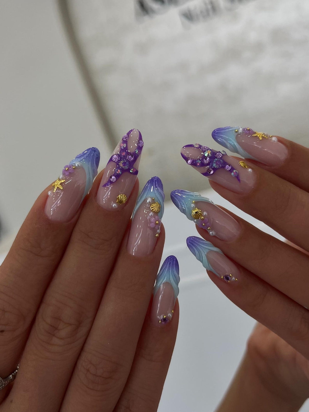 Almond-shaped nails featuring a French manicure base with gradient purple and blue tips. The design includes three-dimensional embellishments such as gold starfish, small seashells, tiny white pearls, and purple rhinestones.