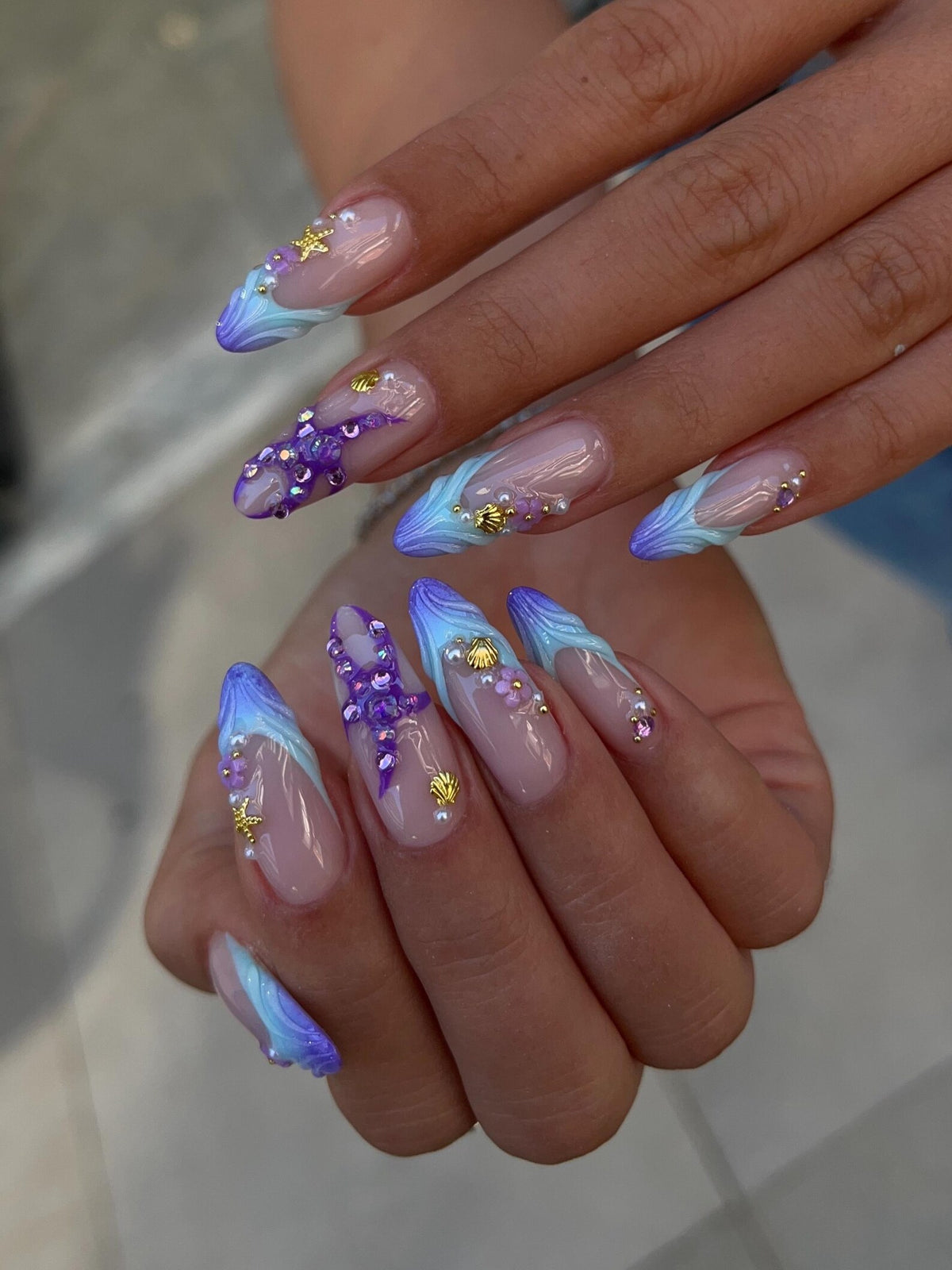 Almond-shaped nails featuring a nude base with blue and purple gradient tips. The design includes 3D embellishments such as gold starfish, gold seashells, small white pearls, and purple crystals arranged in intricate patterns on the nails.