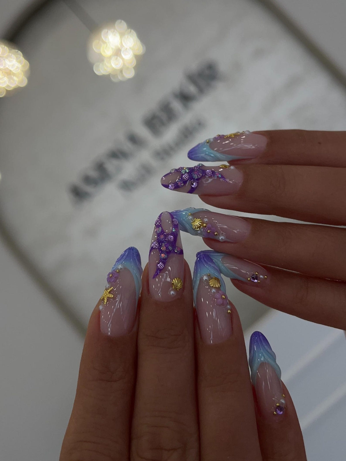 Long almond-shaped nails featuring a French manicure style with blue and purple gradient tips. The nails are decorated with three-dimensional purple starfish, gold seashell charms, small white pearls, and tiny purple floral accents.