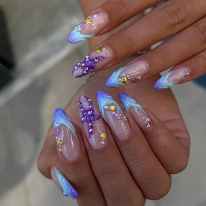 Long almond-shaped nails featuring a French manicure style with blue and purple gradient tips. The nails are decorated with small gold starfish, seashell charms, tiny white pearls, and purple floral accents embedded on the nail surface.