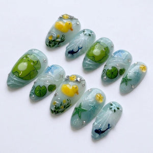 A set of ten press-on nails featuring a light blue and translucent base with aquatic designs including yellow rubber ducks, green frogs on lily pads, dark blue dolphins, green starfish, and small white floral accents with silver rhinestones.