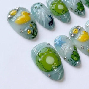 A set of press-on nails featuring a light blue base with hand-painted designs including yellow rubber ducks, green frogs on lily pads, a blue starfish, and small black fish, accented with tiny clear rhinestones and white floral details.