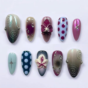 A set of ten almond-shaped press-on nails featuring diverse designs including purple and white orchid 3D floral accents, dark blue and light blue polka dots, silver swirl patterns, metallic pink finishes, and small embedded clear rhinestones.