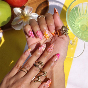 Almond-shaped nails featuring a French manicure base with intricate pink and orange floral art, gold swirls, and small rhinestones. The fingers are adorned with multiple gold rings, including a shell and starfish design.