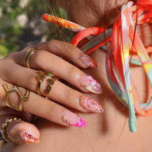 A hand adorned with multiple gold rings, including one with script lettering, and almond-shaped nails featuring intricate pink and orange patterns, gold swirls, and small embedded rhinestones, set against a tie-dye fabric strap.