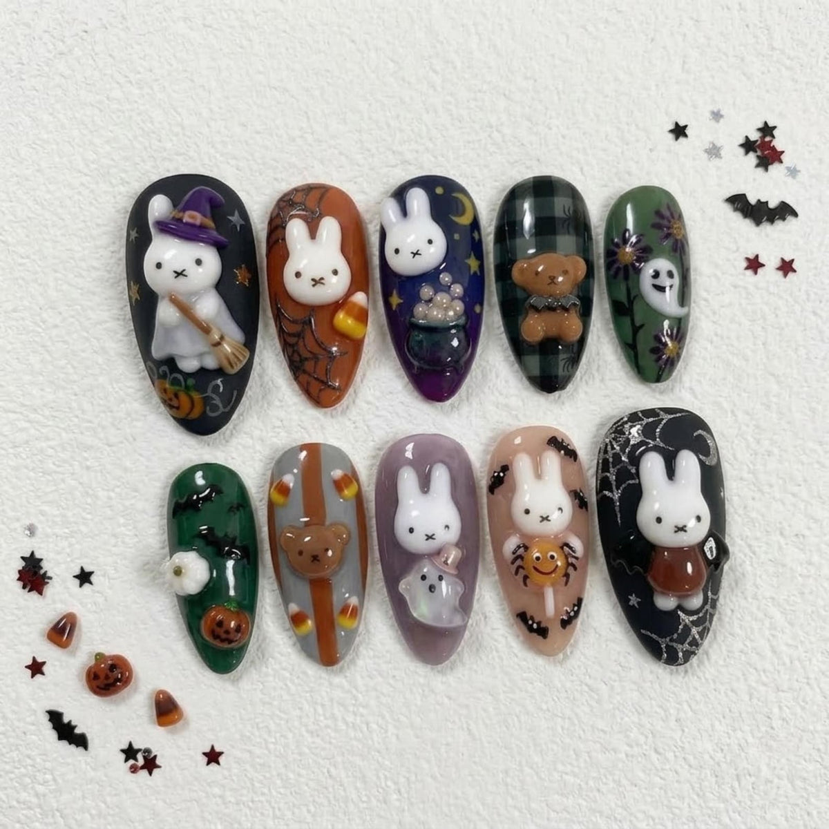 A set of ten almond-shaped press-on nails featuring Halloween-themed designs. The nails display 3D white rabbit figures, pumpkins, ghosts, a teddy bear, candy corn, spider webs, and bats in a color palette of black, orange, green, and purple.