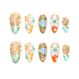 A set of ten almond-shaped press-on nails featuring a nude base with intricate gold line work, light blue floral accents, and small embedded rhinestones. The designs include swirling patterns, layered petals, and metallic gold details.