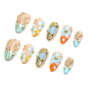 A set of ten press-on nails featuring a mix of nude, light blue, and orange base colors. The designs include raised white floral petals, gold metallic swirls, and small iridescent rhinestones arranged in various decorative patterns.