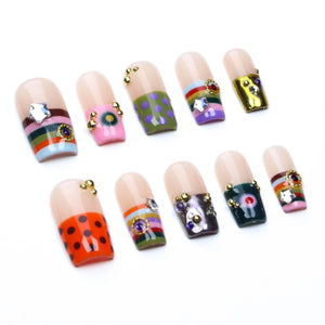 A set of ten press-on nails featuring a nude base with various decorative designs including horizontal stripes, polka dots, and solid colors. Each nail is embellished with small gold-toned metal beads and sparkling rhinestones.