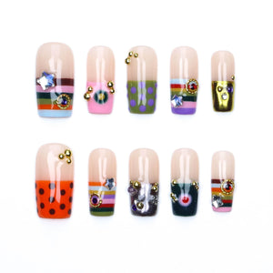 A set of ten press-on nails featuring a nude base with various decorative elements including horizontal stripes, polka dots, metallic gold accents, small colorful rhinestones, and star-shaped charms arranged in two rows of five.