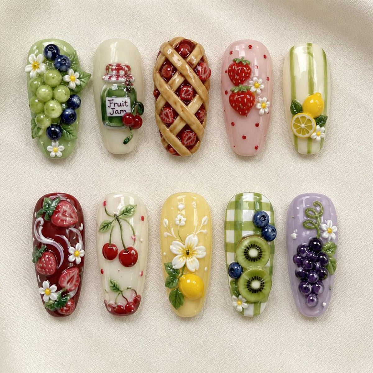 A set of ten press-on nails featuring 3D fruit and dessert designs. Patterns include grapes, a jar labeled Fruit Jam, a lattice pie, strawberries, lemons, cherries, daisies, kiwi slices, and blueberries on various colored backgrounds.