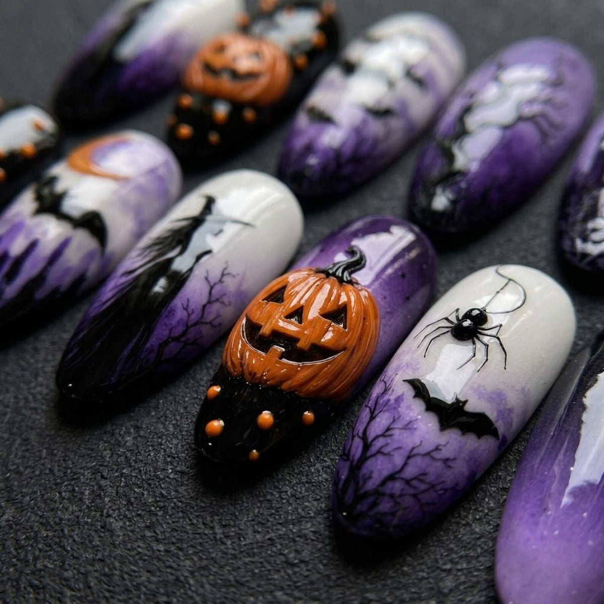 A set of press-on nails featuring Halloween-themed designs including orange 3D pumpkins, black silhouettes of witches, bats, and spiders, set against a gradient background of white and deep purple with painted bare tree branches.