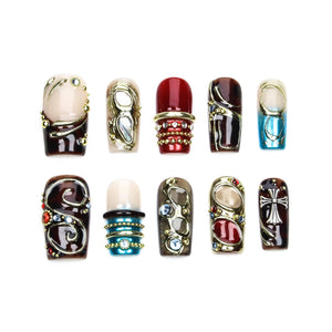 A set of ten press-on nails featuring diverse designs including deep red, brown, and blue tones. The nails are decorated with metallic gold and silver accents, intricate swirls, small embedded rhinestones, and a prominent silver cross emblem.