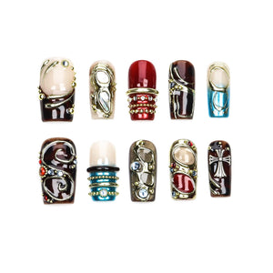 A set of ten press-on nails featuring diverse designs including deep red, brown, and blue tones. The nails are decorated with metallic gold accents, embedded rhinestones, swirling patterns, and a silver cross emblem on one nail.