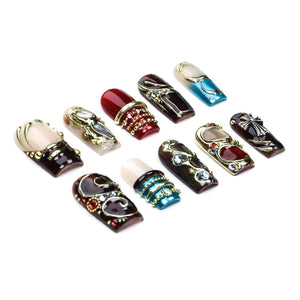 A set of ten press-on nails featuring various designs including deep red, dark brown, and blue base colors. The nails are decorated with gold-toned metal accents, small clear rhinestones, and intricate metallic patterns, including a cross.