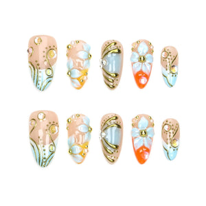 A set of ten almond-shaped press-on nails featuring a nude base with intricate gold swirling lines, small clear rhinestones, and raised white and light blue floral accents. Some nails include vibrant orange tips with white flower designs.