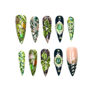 A set of ten stiletto-shaped press-on nails featuring diverse designs including green and brown abstract patterns, black lace textures, raised floral motifs, ornate gold-framed green cabochons, heart charms, and metallic gold accents.