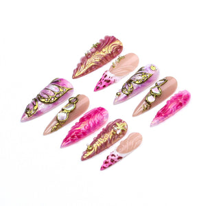 A set of ten press-on nails featuring a pointed stiletto shape. The designs include shades of pink, magenta, and nude, accented with gold metallic swirls, intricate butterfly wing patterns, and small embedded pearls and rhinestones.