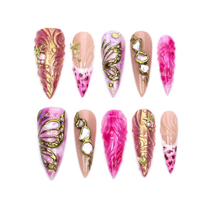 A set of ten stiletto-shaped press-on nails featuring a variety of designs including pink and gold swirling textures, gold butterfly wing outlines, metallic gold accents, small starfish charms, and pink leopard print patterns.